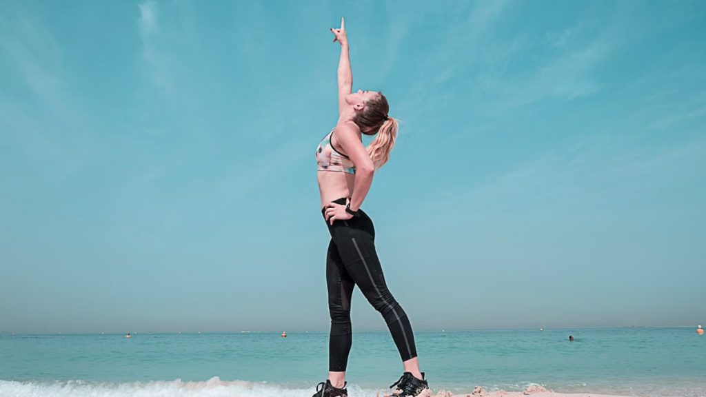 A woman performing a stretching exercise on a sunny beach in Dubai.